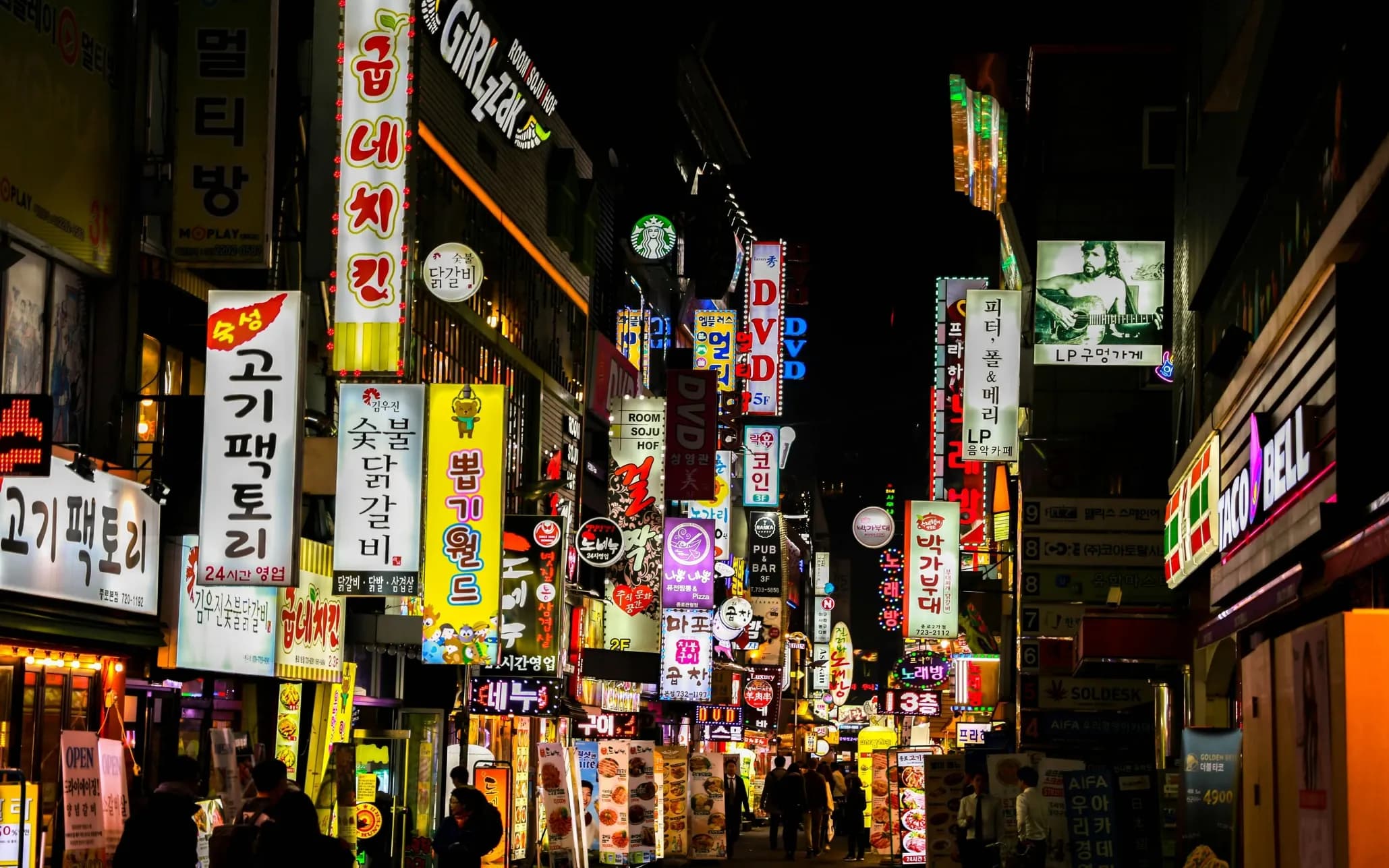 A busy street in South Korea at night filled with colorful neon signs in Korean, restaurants and shops on both sides, and people walking under bright city lights.